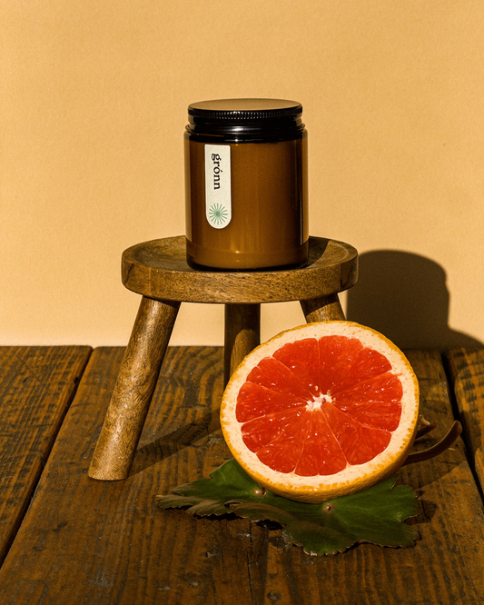 Amber jar of Gronn essential oil candle on a rustic stool, surrounded by fresh grapefruit cut in half, placed on top of geranium leaves—showcasing natural ingredients and eco-friendly design.