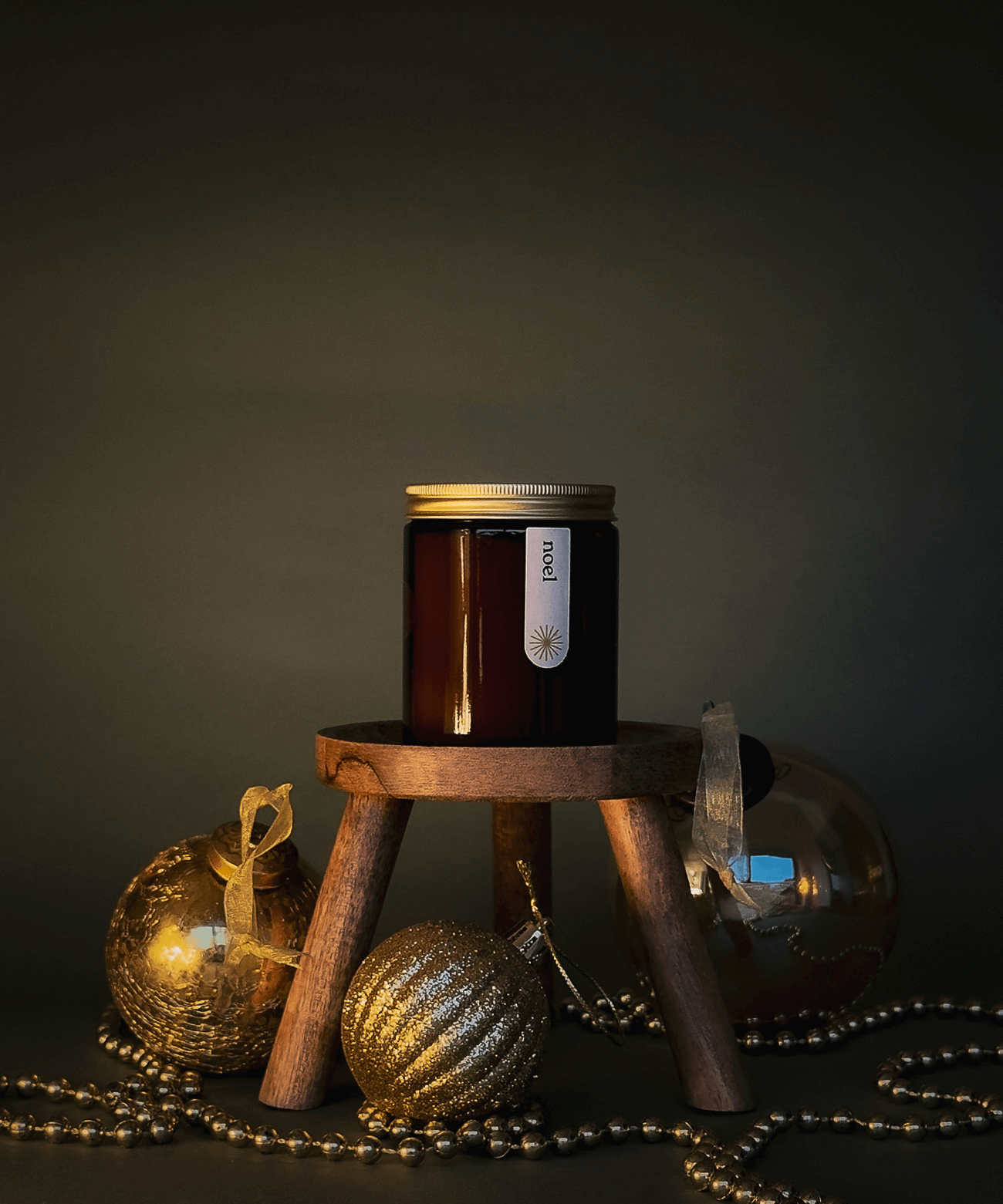 Noel candle on a wooden stand surrounded by gold toned baubles and gold beads.