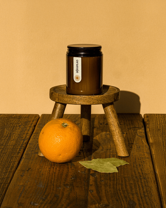 Amber jar of Oransje essential oil candle on a rustic stool, surrounded by fresh orange, and bay leaves—showcasing natural ingredients and eco-friendly design.